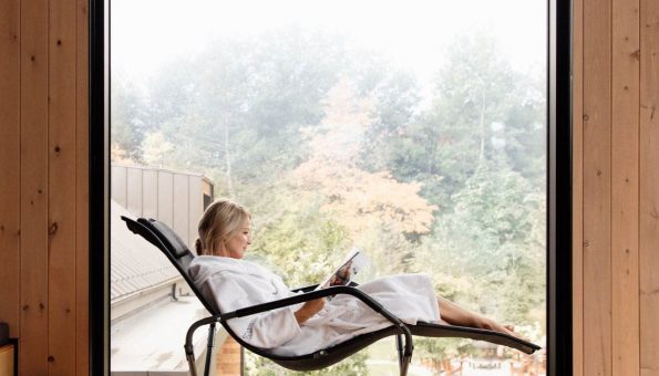 Femme détendue en peignoir, lisant dans un fauteuil inclinable moderne, avec une vue sur un paysage automnal à travers une grande baie vitrée dans un intérieur en bois.