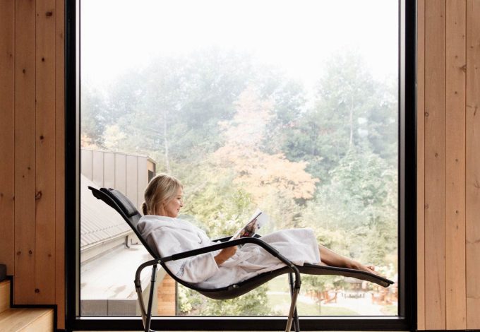 Femme détendue en peignoir, lisant dans un fauteuil inclinable moderne, avec une vue sur un paysage automnal à travers une grande baie vitrée dans un intérieur en bois.