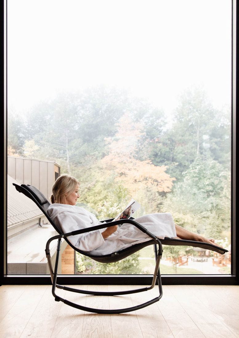 Femme détendue en peignoir, lisant dans un fauteuil inclinable moderne, avec une vue sur un paysage automnal à travers une grande baie vitrée dans un intérieur en bois.